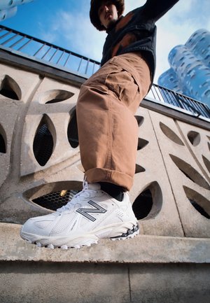 Person trägt weiße New Balance Sneakers und braune Hose und steigt von einem Betonabsatz mit gemusterten Aussparungen vor blauem Himmel herunter.