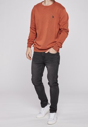 Suéter naranja de canalé con cuello redondo y logotipo negro, combinado con jeans ajustados negros y zapatillas blancas, mostrando un atuendo casual.