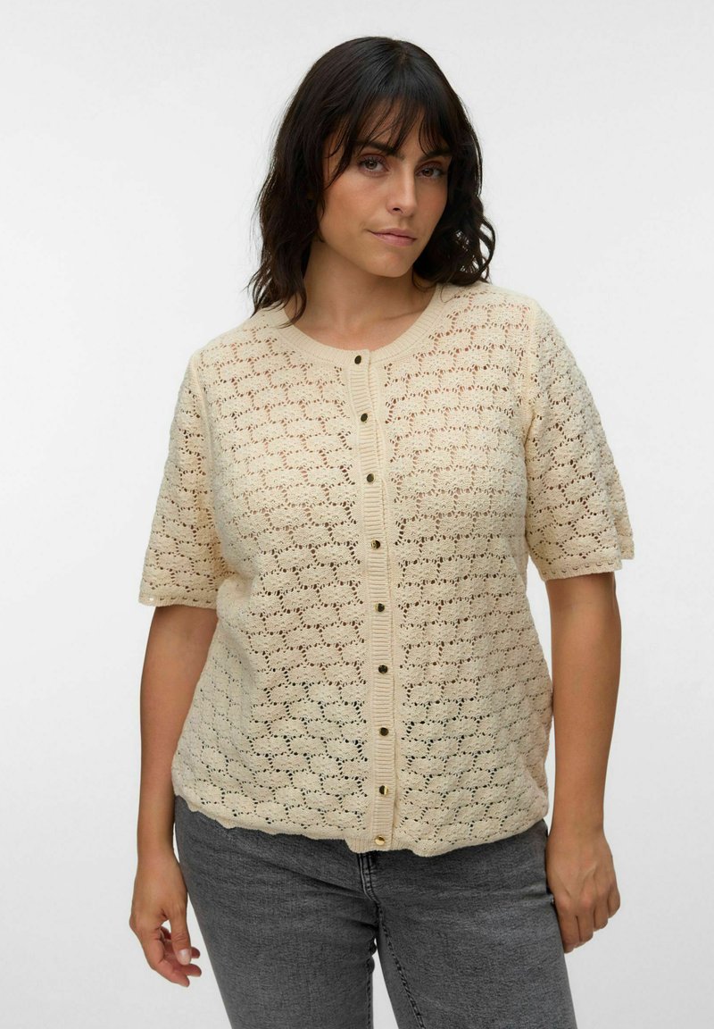 Femme aux cheveux foncés portant un cardigan crocheté beige à manches courtes et boutons et un jean gris sur un fond blanc uni.