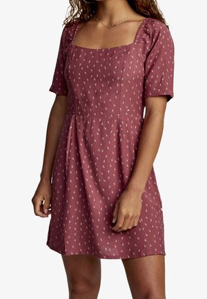 Femme portant une robe mauve à manches courtes avec un petit motif blanc, col carré et taille froncée, montrée des épaules jusqu'au milieu des cuisses.