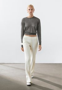 Grauer Strickpullover mit rundem Ausschnitt, kombiniert mit cremefarbenen, strukturierten, weit geschnittenen Hosen mit vertikalen Streifen. Beinhaltet ein Armband als Accessoire.