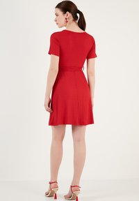 Red ribbed dress with a fitted bodice, short sleeves, and a flared skirt. Features a tie belt accent. Worn with tan and red heeled sandals.