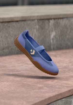 Blå Mary Jane-sneaker med brun sål, gyldent stjerne- og chevronlogo, vist i luften over stensat brostensbelægning.