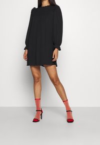 Robe noire à manches longues avec un encolure smockée et des épaules volantes, accompagnée de chaussettes à paillettes rouges et de sandales à talons noires.