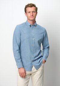 Camicia azzurra chiara con bottoni realizzata in tessuto morbido, dotata di colletto classico, maniche lunghe e un orlo leggermente curvo; indossata con pantaloni bianco sporco.