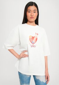Hvit oversized t-skjorte med et fargerikt grafisk motiv av en kvinne med et hjerte-design. Korte ermer og mykt stoff.