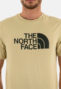 Béžové tričko vyrobené z měkké tkaniny, které má výrazné černé logo s křivkovými prvky, krátké rukávy a kulatý výstřih.