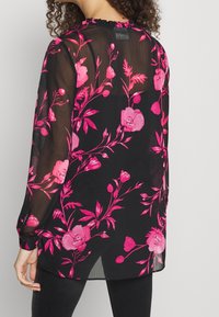 Blusa nera trasparente con maniche lunghe, caratterizzata da un motivo floreale in tonalità di rosa, scollatura tonda con rouches e orlo curvo.