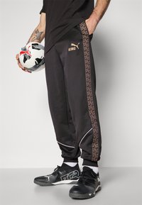 Sorte træningsbukser med mønstrede sidestriber, hvide detaljer og et logo. Personen holder en sort-hvid fodbold. Sorte sneakers er iført.