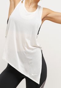 Witte sporttanktop met een asymmetrische zoom en zwarte zijpanelen, met een klein Nike-logo aan de voorkant. Draagt zwarte leggings.