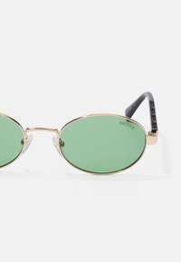 Gold metal-framed sunglasses with green lenses, oval shape, transparent nose pads, and black patterned arms featuring brand logo.