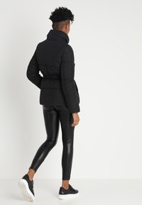 Osoba ubrana w czarną kurtkę puchową, czarne skórzane legginsy i czarne sneakersy, idąca w stronę wyjścia na jednolitym białym tle.