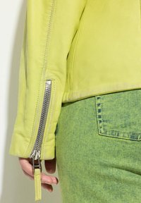 Veste en cuir vert lime avec une fermeture éclair argentée au niveau du poignet, associée à un denim texturé vert assorti. Gros plan sur la manche et la main.