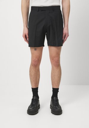 Person iført sorte skræddersyede shorts, sorte strømper og sorte sko, der står mod en ensartet lys baggrund.