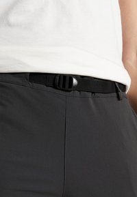 Czarny regulowany pasek wykonany z nylonu, z gładką plastikową klamrą i mocnym materiałowym paskiem, przypięty do szarych spodni.