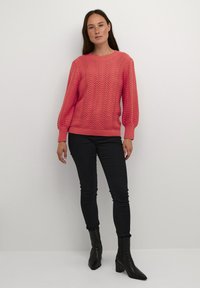 Bright coral knitted sweater with a wave pattern, long sleeves, ribbed cuffs, paired with black skinny jeans and pointed ankle boots.