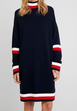 Marineblaues Pulloverkleid mit weißen und roten Streifen am Kragen, an den Bündchen und am Saum, getragen von einer Person mit welligem blondem Haar.