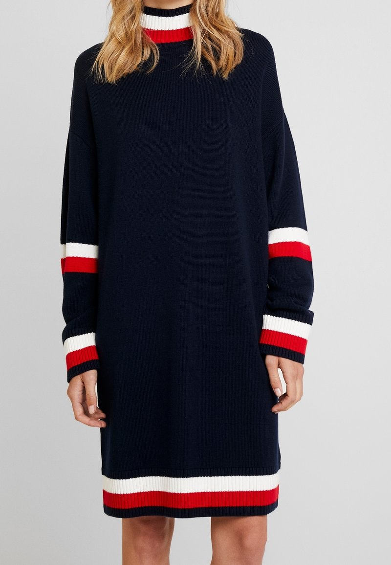 Robe-pull bleu marine avec des rayures blanches et rouges sur le col, les poignets et l’ourlet portée par une personne aux cheveux blonds ondulés.