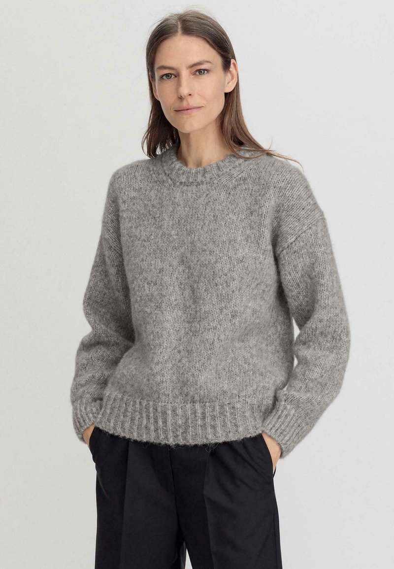 Suéter gris de punto con cuello redondo, hombros caídos y dobladillo acanalado. Textura suave y corte relajado, combinado con pantalones oscuros.