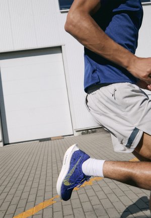 T-shirt de sport bleu, shorts gris clair et chaussures de course violettes avec des accents jaunes ; dessus en maille texturée et semelle blanche rembourrée.