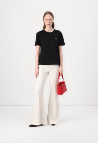 T-shirt en coton noir avec un petit logo doré, associé à un pantalon blanc évasé et un sac à main rouge. Texture lisse et coupe ajustée.