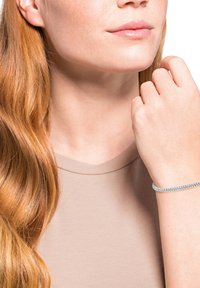 Mujer con cabello largo y rojo, vistiendo una blusa beige y una pulsera de tenis plateada en su muñeca, con la mano cerca de su mentón.