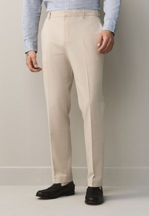 Homme portant un pantalon beige, une chemise claire à motifs et des chaussures noires, debout devant un fond neutre.