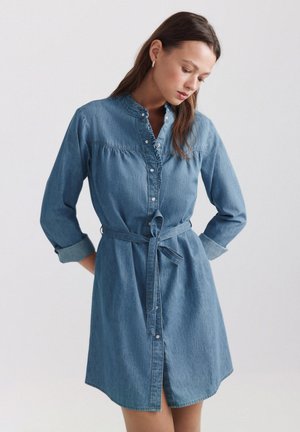 Femme portant une robe en jean bleu boutonnée, manches longues, ceinture à la taille et col à volants, regardant vers le bas avec les mains dans le dos.