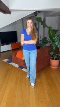 Νεαρή γυναίκα με μπλε πουκάμισο και τζιν στέκεται με σταυρωμένα τα χέρια σε ένα σαλόνι με καφέ καναπέ και μεγάλο φυτευμένο φυτό.