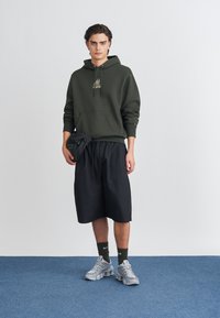 Σκούρο πράσινο φούτερ με κορδόνια, μπροστινή τσέπη, συνδυασμένο με μαύρες βερμούδες μέχρι το γόνατο. Γκρι αθλητικά παπούτσια με λεπτομέρειες από δίχτυ και μαύρες κάλτσες.