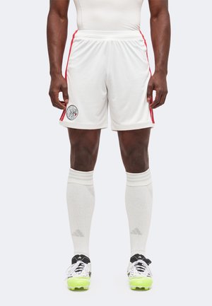 Male athlete wearing white soccer shorts with red stripes, white knee-high socks, and white cleats with neon yellow soles.