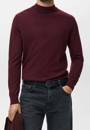 Mand iført en vinrød, langærmet rullekravstrøje, stoppet ned i sorte jeans med et sort bælte, som holder et matchende vinrødt stykke tøj.