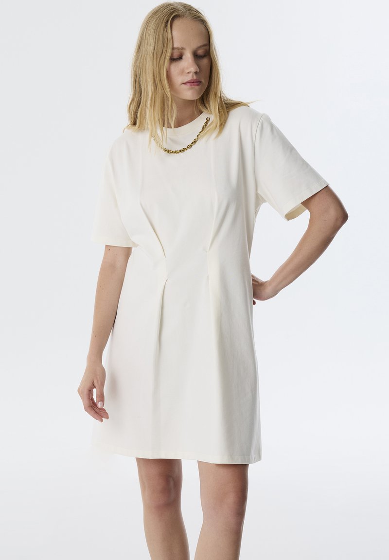 Ein kurzärmliges weißes Kleid mit strukturiertem Finish, seitlichen Falten und Rundhalsausschnitt, ergänzt durch eine goldene Kettenhalskette.