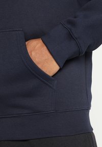 Felpa con cappuccio blu navy con tasca frontale, polsini e orlo a coste. Tessuto morbido con una finitura liscia; presenta una cucitura visibile sulla tasca.