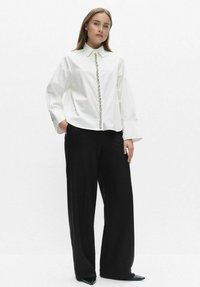 Biała koszula z długim rękawem i szpiczastym kołnierzykiem, ozdobne guziki z przodu, zestawiona z luźnymi czarnymi spodniami i czarnymi szpiczastymi butami.