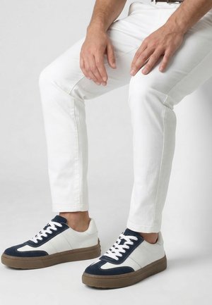 Mann sitzt mit den Händen im Schoß, trägt weiße Hosen und weiße Sneakers mit marineblauen Akzenten und braunen Sohlen.