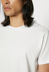 T-shirt en coton blanc avec un col rond, des manches courtes, une texture lisse et un design minimaliste, porté sur le haut du corps d'une personne.