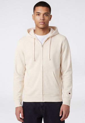 Junger Mann steht, trägt einen hellbeigen Kapuzenpullover mit Reißverschluss und Taschen vorne sowie ein weißes T-Shirt darunter, vor einfarbigem Hintergrund.