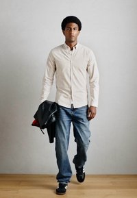 Ανοιχτό μπεζ πουκάμισο με κουμπιά, μπλε τζιν, μαύρα αθλητικά παπούτσια, κρατώντας μία μαύρη δερμάτινη τζακετ. Απλός σχεδιασμός, δομημένη εφαρμογή, χαλαρή εμφάνιση.