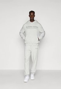 Modello maschile che indossa un hoodie grigio chiaro e pantaloni della tuta abbinati con la scritta "BJÖRN BORG TRAINING CLUB MONACO", in piedi con le mani nelle tasche.