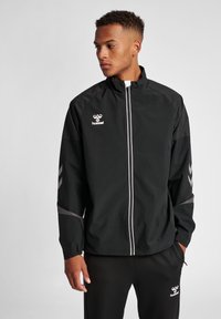 Schwarze, leichte Sportjacke mit hohem Kragen, weißem Reißverschluss und reflektierenden Streifen. Verfügt über ein Logo auf der Brust und Chevron-Muster auf den Ärmeln.