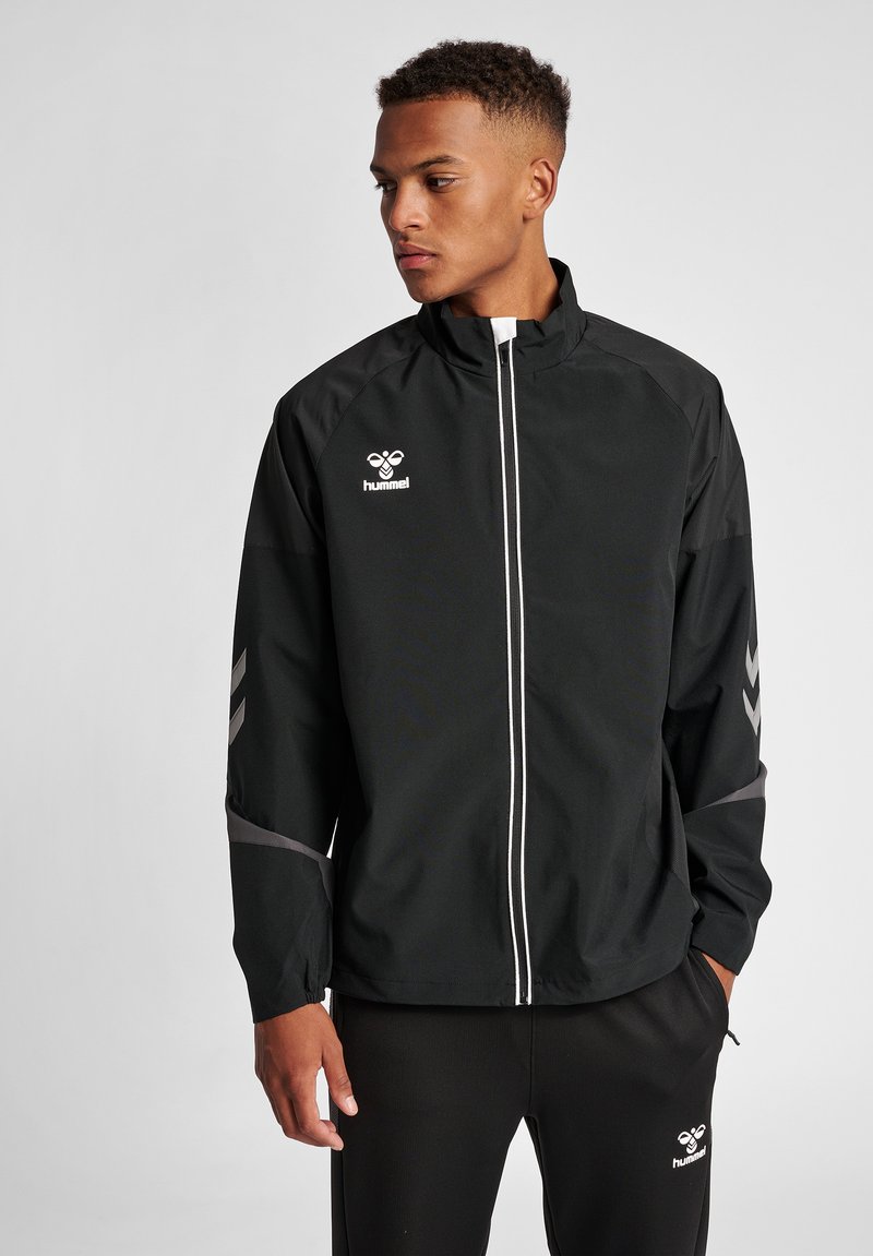 Schwarze, leichte Sportjacke mit hohem Kragen, weißem Reißverschluss und reflektierenden Streifen. Verfügt über ein Logo auf der Brust und Chevron-Muster auf den Ärmeln.
