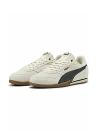 Zapatillas Puma con parte superior de lona color crema, franjas laterales negras, suela de goma texturizada y cordones en la parte delantera. Presenta un logo en la lengüeta.