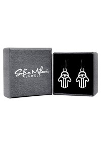 Silverfärgade örhängen formade som en hand med ett öga, visade i en grå texturerad låda med svart inredning och vit logotyp.