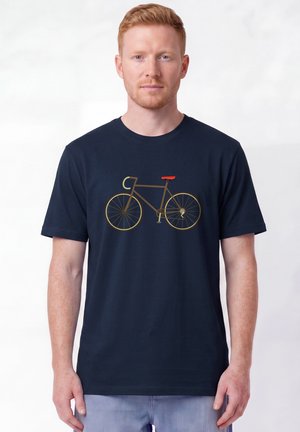 Junger Mann mit heller Haut und roten Haaren, der ein dunkelblaues T-Shirt mit braunem Fahrradmotiv und hellblaue Jeans trägt, steht vor einem einfachen Hintergrund.
