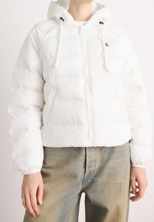 Biała puchowa kurtka z kapturem i troczkami, noszona z wysokim stanem i wypłowiałymi niebieskimi dżinsami, z zamkiem zapiętym, ręce swobodnie opuszczone wzdłuż ciała.