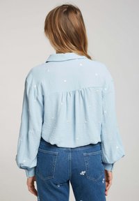 Blouse bleu clair en crêpe avec de longues manches bouffantes et une petite broderie florale. Elle présente un col et un design de bas arrière plus long.