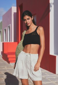 Cropped top nero a maglia con un motivo testurizzato, abbinato a pantaloni corti in lino beige con cordoncino. Il modello porta una borsa intrecciata verde.