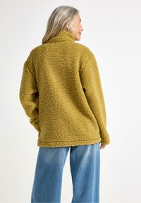 Una donna con lunghi capelli grigi, sorridente mentre indossa una comoda giacca in pile verde oliva e jeans azzurri chiari, rivolta leggermente a sinistra.