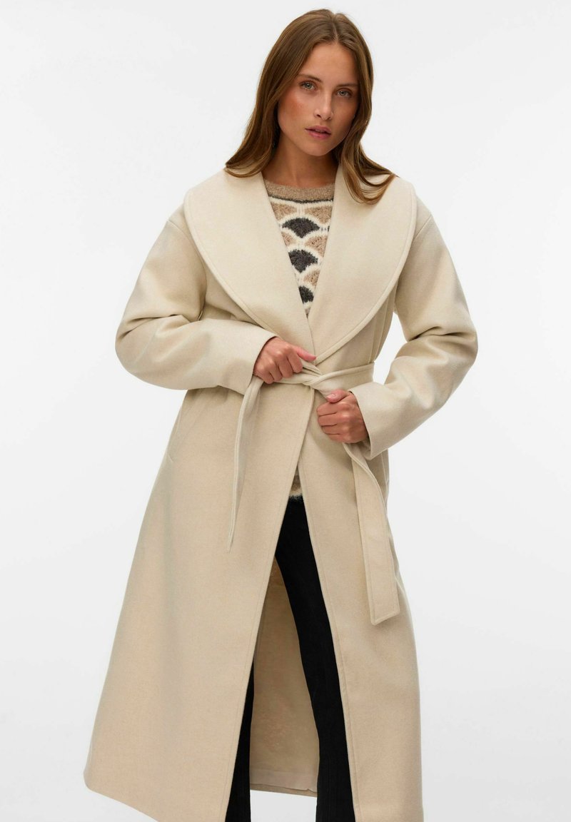 Manteau en laine beige avec un grand col, des manches longues et une taille nouée. Le manteau est porté sur un pull à motifs et présente une coupe droite.